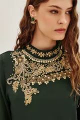 Camisa de viscosa color verde oscuro, con cuello alto y mangas tres cuartos terminadas en volados. Presenta un elaborado bordado artesanal en el pecho con pedrería dorada y detalles decorativos en la cintura.