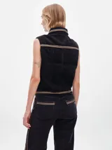 Chaleco de pana negro con cierre frontal de botones metálicos dorados. Presenta detalles de costuras en contraste en el cuello, cartera y canesú, e incluye un pequeño parche con forma de bandera en el pecho.