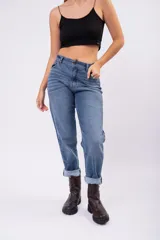 Pantalón tipo mom jean de tiro alto, confeccionado en denim rígido de color azul medio con efecto lavado. Presenta un corte recto y holgado con ruedos doblados, cierre frontal con botón y bolsillos laterales.