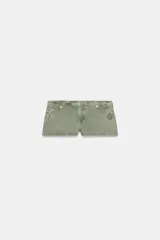 Short mini de tiro bajo en color verde oliva, confeccionado en tejido de algodón con efecto desgastado. Presenta detalles de costuras decorativas, aplicaciones circulares en los laterales y botones metálicos en la cintura. Cuenta con cierre frontal oculto.