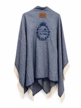 Ruana de tejido de punto con diseño a rayas diagonales en tonos azul y blanco, con flecos en el ruedo y bordado central del escudo nacional. Incluye etiqueta de cuero con marca Bagual.