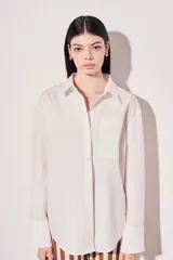 Camisa blanca oversize de algodón, con cuello clásico, cierre frontal con botones de nácar y bolsillo en el pecho.