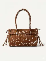 Cartera de tamaño medio confeccionada en cuero vacuno con textura de pelo y estampado animal print tipo vaca. Presenta estructura rígida, doble asa de hombro, cierre superior, detalle de tira horizontal con tachas y hebillas metálicas, y lazos decorativos en los laterales.