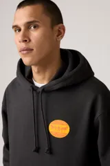Hoodie negro de corte relajado con capucha y bolsillo canguro frontal. Presenta un parche o estampado circular en color naranja y amarillo en el pecho.