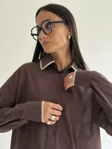 Camisa oversize de color marrón con cuello camisero y puños con detalle de bordado festoneado en color blanco. Presenta cierre frontal de botones y mangas largas.