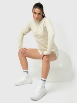 Remera deportiva de manga larga en color beige, confeccionada en tela reciclada suave. Presenta un diseño con cuello alto y medio cierre frontal, además de un bolsillo lateral integrado. Las mangas incluyen aberturas para los pulgares.