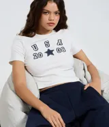 Remera baby look color blanco marfil de algodón con elastano, corte ajustado, cuello redondo, mangas cortas y estampa frontal estilo universitario con el texto "USA 2008" y una estrella en azul marino.