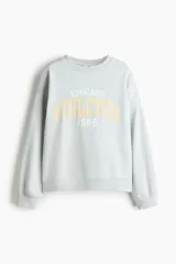 Buzo oversize celeste de cuello redondo, con hombros caídos, puños y bajo acanalados, y diseño de texto frontal con la inscripción "CHICAGO ATHLETICS 1985".