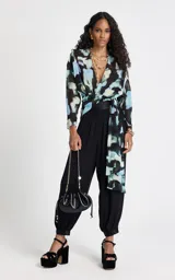 Blusa de tejido fluido con estampado abstracto en tonos azules, negros y beige. Presenta un diseño cruzado con escote en V, mangas 3/4 anchas y un lazo lateral ajustable que define la silueta.