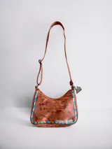 Cartera y bandolera de cuero color marrón tostado con efecto desgastado, detalles de tachas y costuras decorativas en tonos turquesa y marrón, y correa regulable.