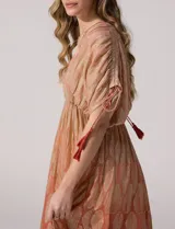 Vestido largo de gasa color salmón con un sutil patrón estampado brillante en tono dorado. Cuenta con escote en V cruzado, cintura imperio elastizada y mangas cortas con tiras ajustables terminadas en borlas.