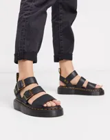 Sandalia Dr. Martens Gryphon Quad negra con plataforma, fabricada con piel Pisa. Presenta tres correas ajustables con hebillas de latón, pespuntes amarillos distintivos en la suela y tirador trasero con el logo AirWair.