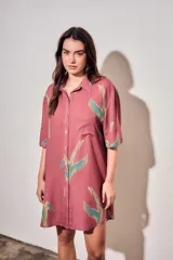 Vestido camisero corto de color rosado con estampado de aves abstractas en tonos verdes y azules. Cuenta con cuello camisero, cierre frontal con botones, mangas cortas y un bolsillo aplicado en el pecho.