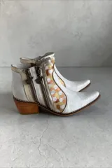Botas de caña corta de cuero vacuno en tono blanco y detalles metalizados, con diseño pintado a mano en tonos dorados, blancos y terracota en los laterales. Posee taco cuadrado de madera, punta fina y doble cierre lateral con tiradores metálicos personalizados.