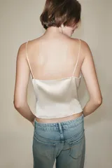 Top crop satinado de color crudo, con escote en pico, tirantes finos y detalle de encaje combinado en el escote y al frente. Presenta cierre frontal con pequeños botones y bajo de corte asimétrico.