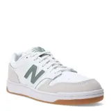 Championes urbanos New Balance Court, color blanco con detalles en verde y gris, confeccionados en cuero y gamuza, con logo "N" característico en los laterales y suela de goma color caramelo.
