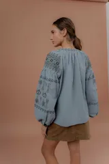 Blusa de estilo bohemio color celeste, con mangas abullonadas y amplias, escote redondo con tiras colgantes y borlas, y detalles de bordados geométricos en contraste de color en el pecho y las mangas.