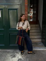 Cartera tipo tote de cuero con diseño de franjas verticales multicolores en tonos tierra, rojo y azul. Cuenta con correas trenzadas de cuero y cierre superior.