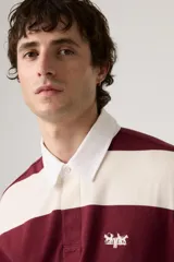 Remera tipo rugby de manga larga con diseño de rayas horizontales anchas en color bordó y crema. Presenta cuello polo blanco, cierre frontal con botones ocultos y un pequeño logo bordado en el pecho.