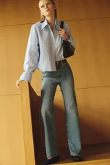 Camisa de manga larga color celeste con finas rayas verticales blancas y amarillas. Presenta cuello clásico, cierre frontal con botones y bajo redondeado con espalda ligeramente más larga.