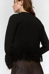 Chaqueta de punto color verde oscuro, con cuello redondo, manga larga y cierre frontal con un solo botón. Presenta detalles de flecos en el ruedo y en los puños.