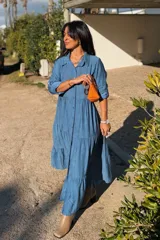 Vestido largo de estilo camisero en color azul, con cuello clásico, cierre frontal de botones y mangas tres cuartos con trabillas. Presenta un diseño de falda con cortes horizontales que le otorgan volumen y movimiento.