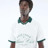 Camiseta tipo polo de manga corta, color blanco con cuello y ribetes en las mangas de color verde oscuro. Presenta un diseño gráfico estampado en el pecho con inscripciones y detalles en tono verde.