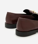 Mocasines de mujer confeccionados en poliuretano color marrón oscuro, con acabado brillante y detalle decorativo metálico dorado en el empeine. Presentan una silueta clásica de punta redondeada y suela plana con pequeño tacón.