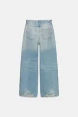 Jeans de tiro bajo y corte wide leg, confeccionados en denim de color celeste claro con lavado desgastado. Presentan detalles de roturas y aplicación de tachas metálicas en los laterales y bolsillos delanteros. Cierre frontal con cremallera y botón.