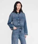 Chaqueta corta de denim azul con cuello camisero de cuero negro y cierre frontal de botones metálicos. Presenta dos bolsillos delanteros con solapa y hombros caídos.