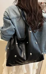 Cartera tipo hobo de cuero sintético negro, con diseño de tachas metálicas plateadas distribuidas por toda la superficie. Cuenta con una correa ancha integrada para llevar al hombro.