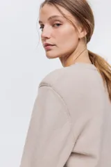 Buzo en punto fino suave de color beige con hombreras y ribete acanalado en cuello redondo, puños y bajo.