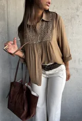 Camisa de color camel con cuello clásico, manga larga y detalle de bordado geométrico en color blanco en la parte frontal superior. Presenta cierre frontal con botones y un corte holgado.