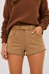 Short de efecto gamuza color camel, de tiro medio, con detalle de tachas metálicas doradas envejecidas en los bolsillos y en el ruedo. Incluye cinturón a tono con hebilla metálica.