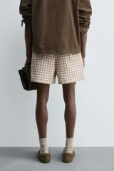 Bermuda de hombre, corte relajado, color beige con estampado de cuadros en tonos marrones y rosados. Presenta cintura elástica, bolsillos frontales y un bolsillo trasero de plastrón con cierre de botón.
