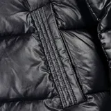 Campera puffer negra, acolchada y liviana, con capucha, cuello alto, cierre frontal con cremallera y botones a presión, dos bolsillos externos y sistema de ajuste regulable en la parte baja.