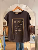 Remera de algodón color beige con corte relajado y diseño asimétrico, más larga en la parte posterior. Presenta un estampado frontal rectangular con letras doradas, una pluma ilustrada y detalles de pedrería decorativa.