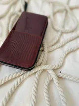 Billetera grande de cuero marca Rupia, con diseño bicolor en tono bordó. Presenta una textura granulada en la parte superior y una textura tipo reptil en la parte inferior. Incluye cierre metálico con tirador de cuero negro y un dije metálico circular plateado con el logo de la marca.