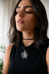 Collar con dije de acero quirúrgico en forma de flor tipo margarita, montado sobre un tiento de cuero negro.