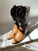 Bota tejana de cuero color marrón claro con puntera fina, caña media con bordados en negro y blanco, y taco bajo.