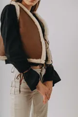 Chaleco de cuero marrón con interior y detalles de corderito sintético blanco, diseño sin mangas con cordones laterales ajustables.