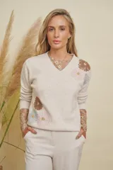 Buzo color beige con cuello en V y mangas largas. Presenta apliques de flores bordadas en tonos marrón, rosa y celeste, con lentejuelas doradas en el centro. Las mangas tienen un detalle de encaje beige que sobresale de los puños.