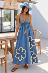 Vestido largo de denim azul oscuro con tirantes finos, escote en V y corte holgado. Presenta bordados ornamentales en el pecho y grandes apliques florales con flecos y perlas en la falda, además de un cordón ajustable en la cintura con borlas colgantes.