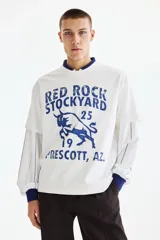 Remera de algodón blanca con estampado azul estilo universitario de toro y texto "Red Rock Stockyard". Presenta cuello redondo, mangas cortas y hombros caídos.
