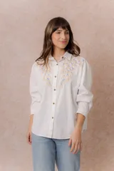Camisa de denim azul con bordados florales en tonos amarillos y celestes sobre los hombros y el pecho. Presenta cuello clásico, cierre frontal con botones y mangas largas con puños.
