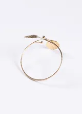 Brazalete abierto para brazo en tono dorado, con diseño entrelazado y dos dijes en forma de concha marina en los extremos.