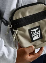 Bolso tipo morral pequeño de la marca Obey, confeccionado en lona de poliéster. Presenta un diseño bicolor en tonos beige y negro, con un parche frontal del logo de la marca en PVC. Cuenta con cierre metálico, bolsillo interno, correa regulable y herrajes plásticos.