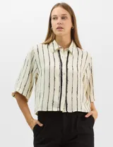 Camisa de corte holgado y silueta recta, con estampado de rayas verticales irregulares en color negro sobre fondo crema. Presenta cuello camisero clásico, mangas cortas amplias y cierre frontal con botones.
