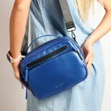 Cartera bandolera de diseño rectangular con acabado metalizado en color azul. Cuenta con un compartimento principal con cierre, un bolsillo frontal con cierre, asa superior fija y correa larga ajustable y extraíble. Incluye el logo de la marca grabado en la parte inferior frontal.