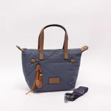 Bandolera tipo shopper azul con diseño acolchado, doble asa de mano en tono tostado y correa ajustable desmontable. Cuenta con cierre superior, bolsillo frontal con cierre y detalle de llavero colgante con cordones naranjas y cuentas.